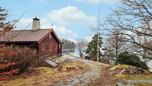 A rustic wooden building with a sloped roof and a chimney, situated by a stone pathway leading to a body of water. - Cozy home in Fjärås with house sea view (Fjärås)