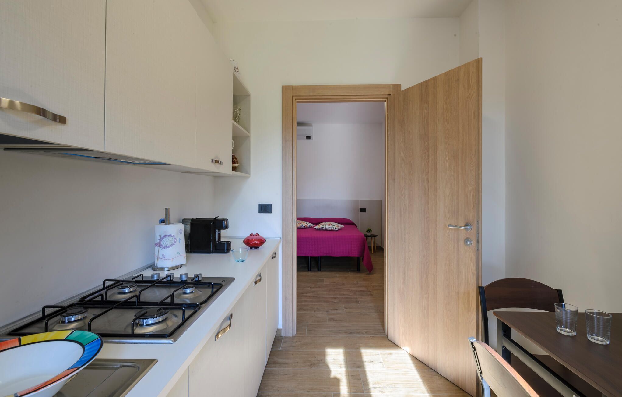 Cocina compacta con estufa a gas, cafetera y una pequeña mesa de comedor con dos vasos. Un pasillo estrecho da a un dormitorio con cama y mesita de noche.
