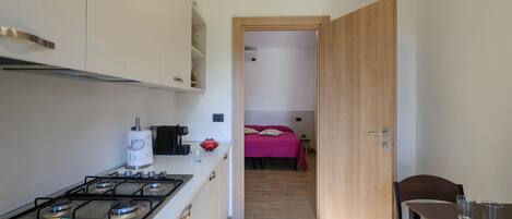 Cocina compacta con estufa a gas, cafetera y una pequeña mesa de comedor con dos vasos. Un pasillo estrecho da a un dormitorio con cama y mesita de noche.