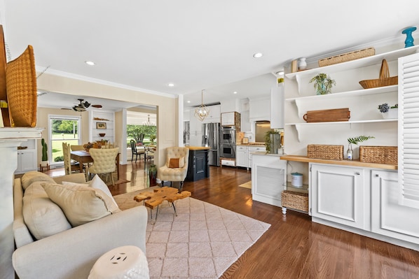 A spacious living room with a large sofa, a dining area with chairs, and a kitchen with white cabinets and wooden shelves.