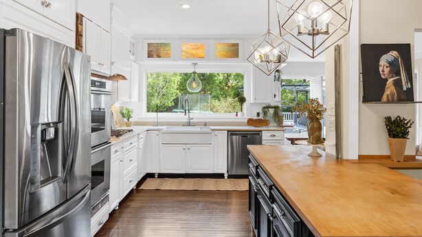 A modern kitchen with white cabinetry, stainless steel appliances, a wooden island, and a view of greenery outside.