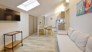 Ein kompakter Wohnbereich mit einer Couch, einem Esstisch mit Stühlen, einem an der Wand befestigten Fernseher, einer Küchenzeile und einem Oberlicht.