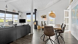 A modern living room with a large gray sectional sofa, a wooden dining table with six chairs, a fireplace, and a large window offering a view of the outdoors. - Beautiful home in Hejls with WiFi (Hejls)