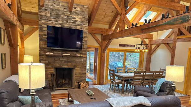 A cozy living room with a stone fireplace, wooden beams, a flat-screen TV, and a dining area with a table and chairs. - New Spacious Acadia Timberframe, Close to Swimming, Central to Hiking and Dining (Mount Desert)