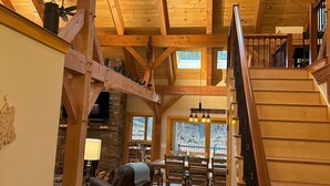 A spacious interior with a high ceiling, wooden beams, and skylights. A staircase leads to an upper level, and a fireplace is visible in the background. - New Spacious Acadia Timberframe, Close to Swimming, Central to Hiking and Dining (Mount Desert)