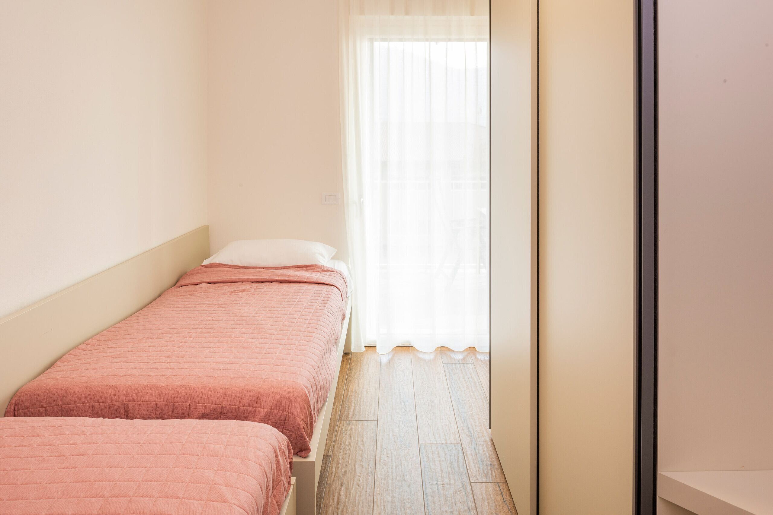 Ein kleiner, schmales Zimmer mit zwei Betten, einem Fenster mit durchsichtigen Vorhängen und einem Holzboden.
