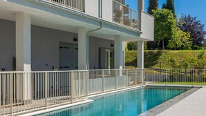 Une piscine entourée d’une clôture blanche, un espace abrité avec des portes, et une vue sur des arbres et une colline.