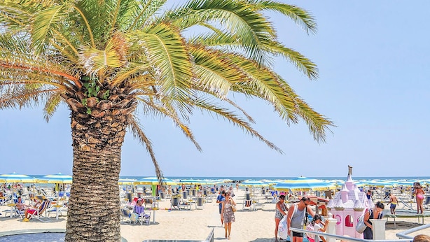 A beach with palm trees, beach chairs, umbrellas, and a castle-like structure.