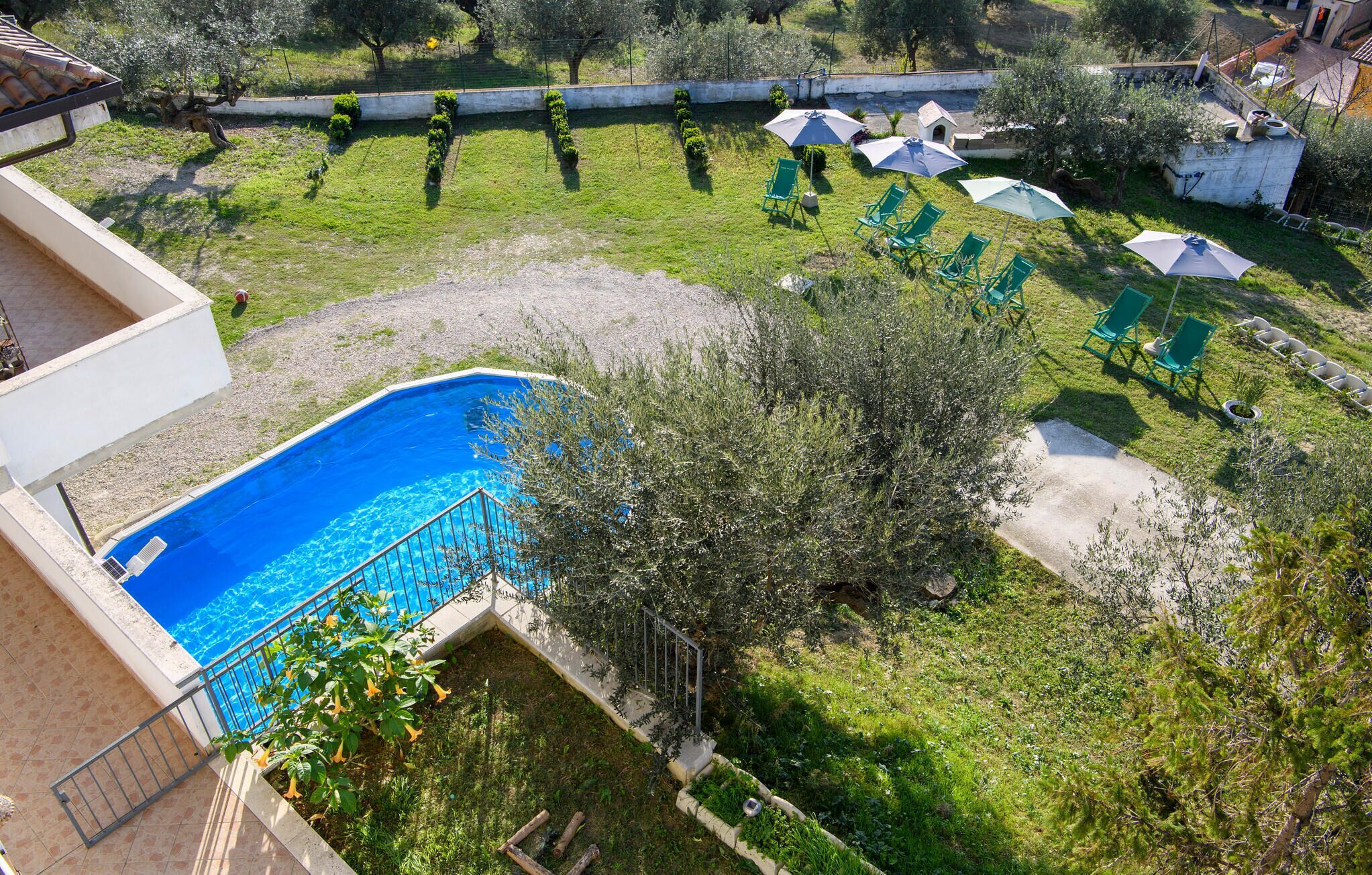Piscina rodeada por um jardim com cadeiras e guarda-sóis verdes, uma pequena casa, e vista para a paisagem ao redor.