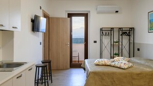 A bedroom with a bed, a TV mounted on the wall, a wooden door leading to a balcony, and a kitchenette with a sink and stools.