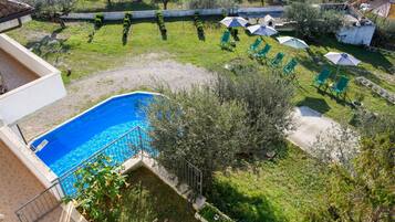 Una piscina circondata da un giardino con sedie e ombrelloni verdi, una piccola casa e vista sul paesaggio circostante.