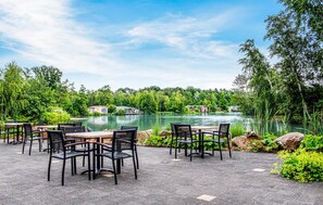 An outdoor seating area with tables and chairs facing a lake, surrounded by greenery and houses in the background. - Lovely home in Oosterwolde with WiFi (Oosterwolde)