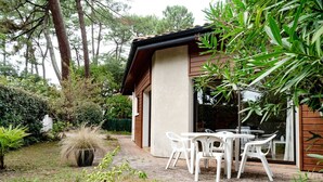 A patio with white plastic chairs and a table, surrounded by greenery and a house with a brown roof. - Capbreton Villa au Domaine de la Cigalière Pour 4 Personnes (CAPBRETON)