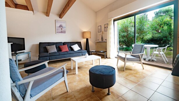 A modern living room with a sofa, coffee table, and chairs. There is a television, a lamp, and a window with a view of greenery. - Capbreton Villa au Domaine de la Cigalière Pour 4 Personnes (CAPBRETON)
