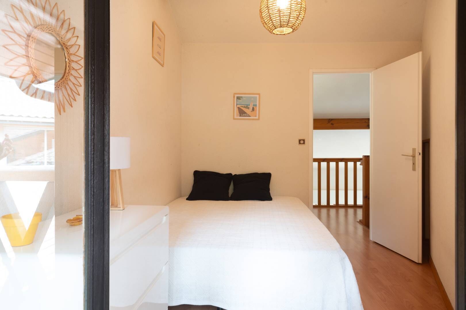Un dormitorio con cama, una mesita de noche con lámpara, un cuadro en la pared y una puerta que da a otra habitación.