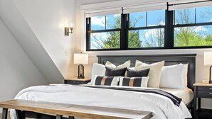 A bedroom with a large bed, two bedside tables, a bench, and a window with a view of trees. - Luxury Ski Chalet · Hot Tub + Sauna + Game Room (Bethel)
