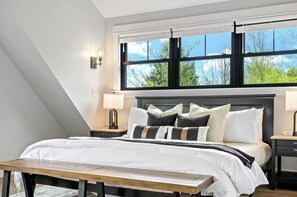 Un dormitorio con una cama grande, dos mesitas de noche, un banco y una ventana con vistas a árboles.