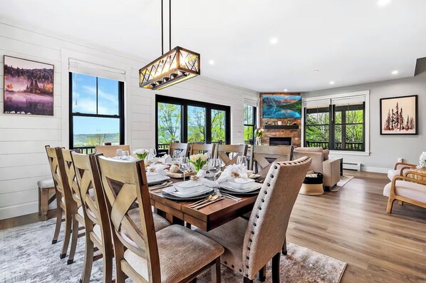 A dining area with a wooden table set for six, surrounded by wooden chairs. There is a large window with a view of greenery outside. - Luxury Ski Chalet · Hot Tub + Sauna + Game Room (Bethel)