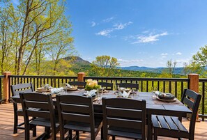 Un área de comedor al aire libre con una mesa dispuesta para cuatro personas, rodeada de sillas, sobre una terraza de madera con vistas a un paisaje montañoso.