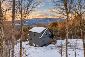 A two-story cabin with a snow-covered roof, surrounded by trees and a mountainous backdrop.