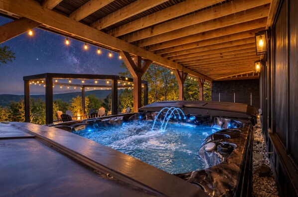 A hot tub area with a wooden pergola and string lights, surrounded by a wooden deck and a view of the mountains.