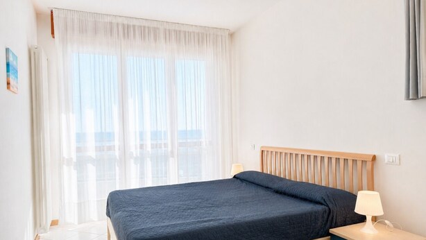 Een hotelkamer met een bed, nachtkastje, lamp en een raam met dunne gordijnen.