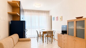 Een woonkamer met een bank, een eettafel met stoelen, een televisie op een kast en een raam met dunne gordijnen.