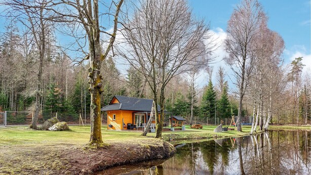 Eine Holzhütte mit geneigtem Dach, umgeben von einem eingezäunten Garten, einem kleinen Teich und Bäumen.
