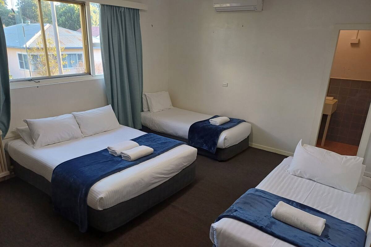 A hotel room with two beds, a window with curtains, a wall-mounted air conditioner, and a view of a house outside.