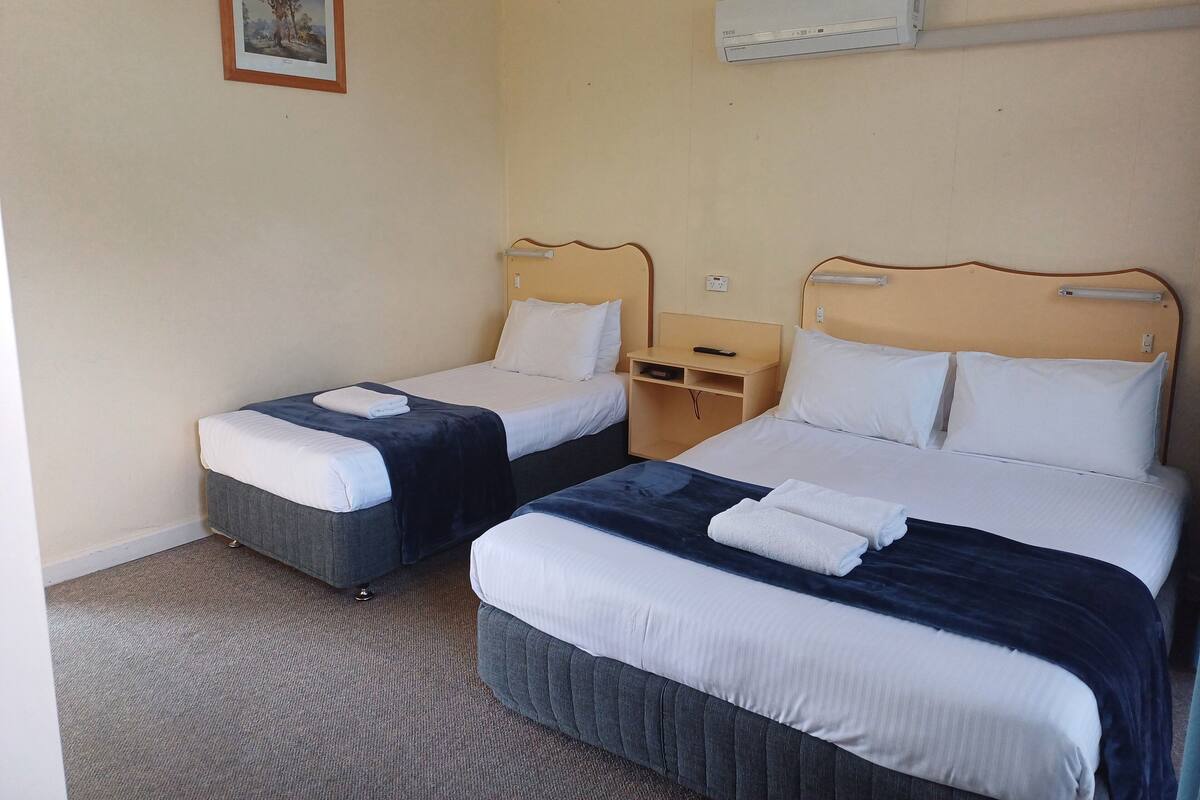 A hotel room with two beds, a small bedside table with a remote control, a wall-mounted air conditioner, and a framed picture on the wall.