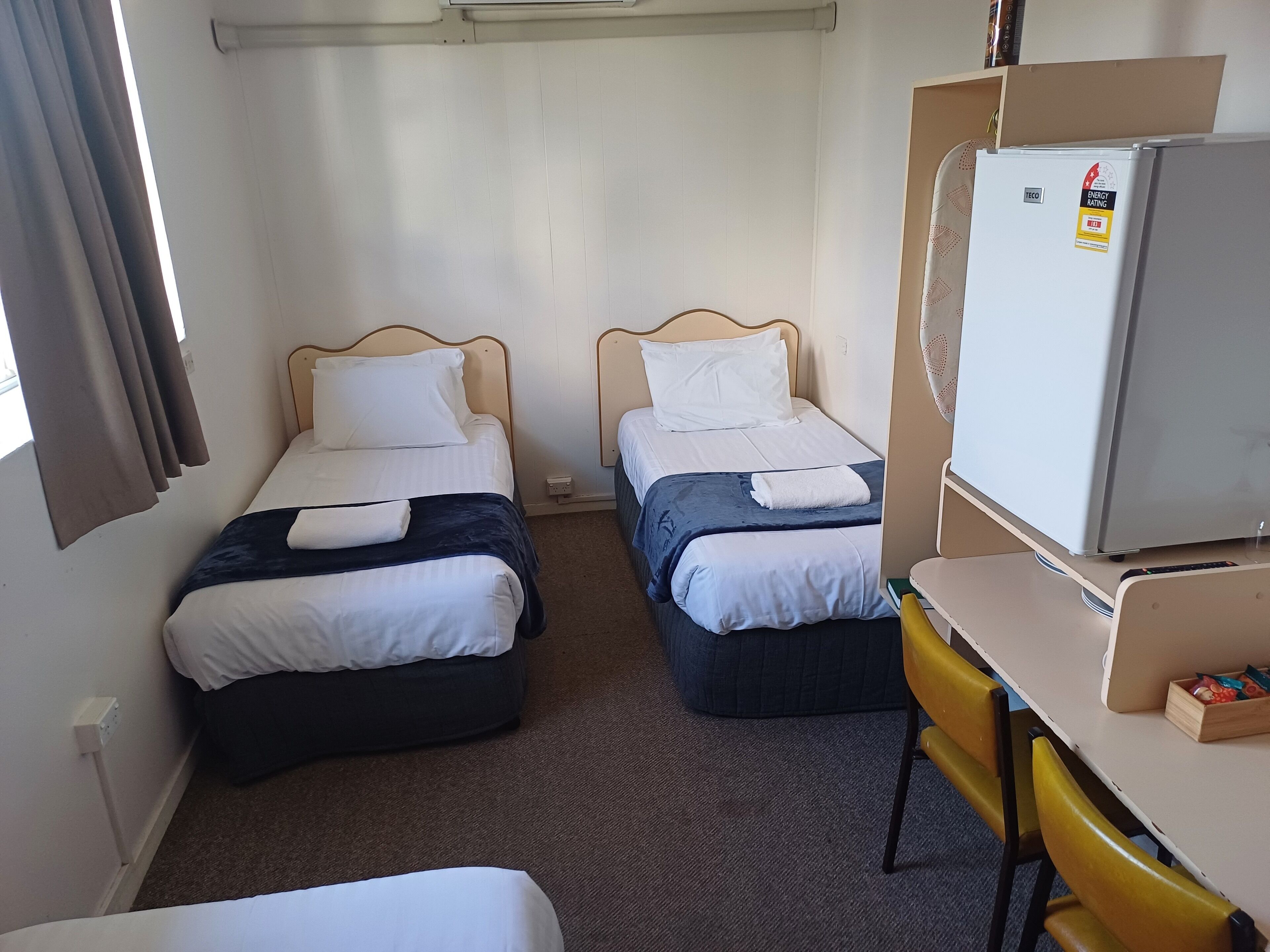 Una habitación de hotel con dos camas individuales, una mesita con una silla, un refrigerador y una ventana con cortinas.