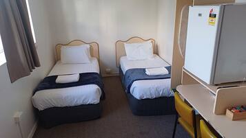 Una habitación de hotel con dos camas individuales, una mesita con una silla, un refrigerador y una ventana con cortinas.