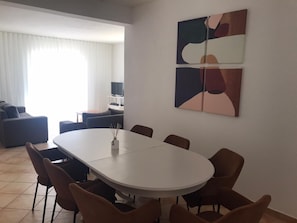 A modern living room with a dining area, featuring a white table and brown chairs, a sofa, and a TV on a stand.