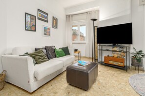 Ein Wohnzimmer mit einem weißen Sofa, einem braunen Hocker, einem Flachbildfernseher, einem Bücherregal und einem Fenster mit Vorhängen.