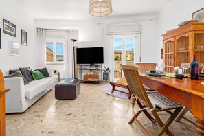 Ein Wohnzimmer mit Sofa, Hocker, Fernseher, Esstisch aus Holz, Stühlen und Bücherregal.