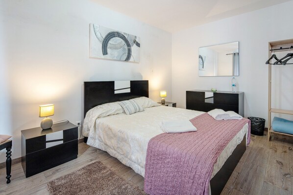 Un dormitorio con cama, mesitas de noche, espejo, una obra de arte en la pared, perchero y suelo de madera.