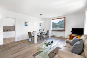 Un salon moderne avec une table basse en verre, un canapé, un coin repas avec des chaises, un téléviseur et une vue sur les montagnes.