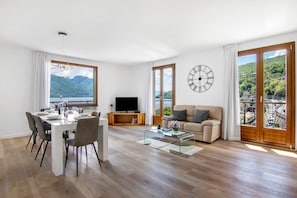 A modern living room with a dining area, a sofa, a TV, and a large window with a mountain view.