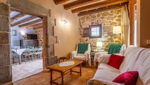 A cozy living room with stone walls, a wooden ceiling, a white sofa with red and green cushions, and a small table with a lace doily.