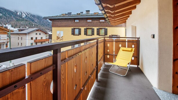 Ein Balkon mit einem gelben Stuhl, Holellentrailering und Blick auf Gebäude und Berge.