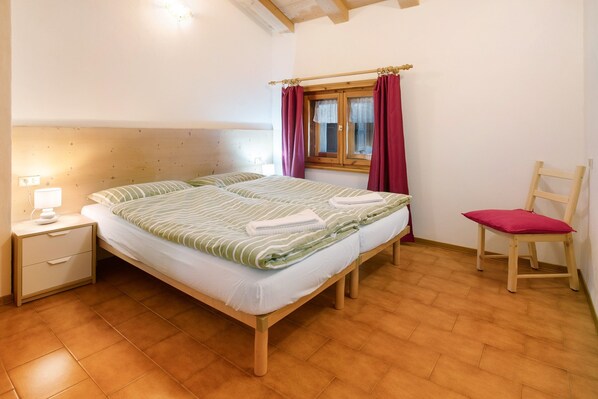 Quarto com cama, mesa de cabeceira, cadeira, janela com cortinas e teto de madeira.