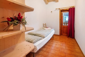 A small room with a bed, a wooden shelf with a vase of flowers, a wooden desk, and a door with a red curtain.