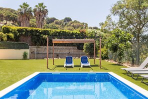 A swimming pool with two lounge chairs under a pergola, surrounded by a well-maintained lawn and a variety of trees.