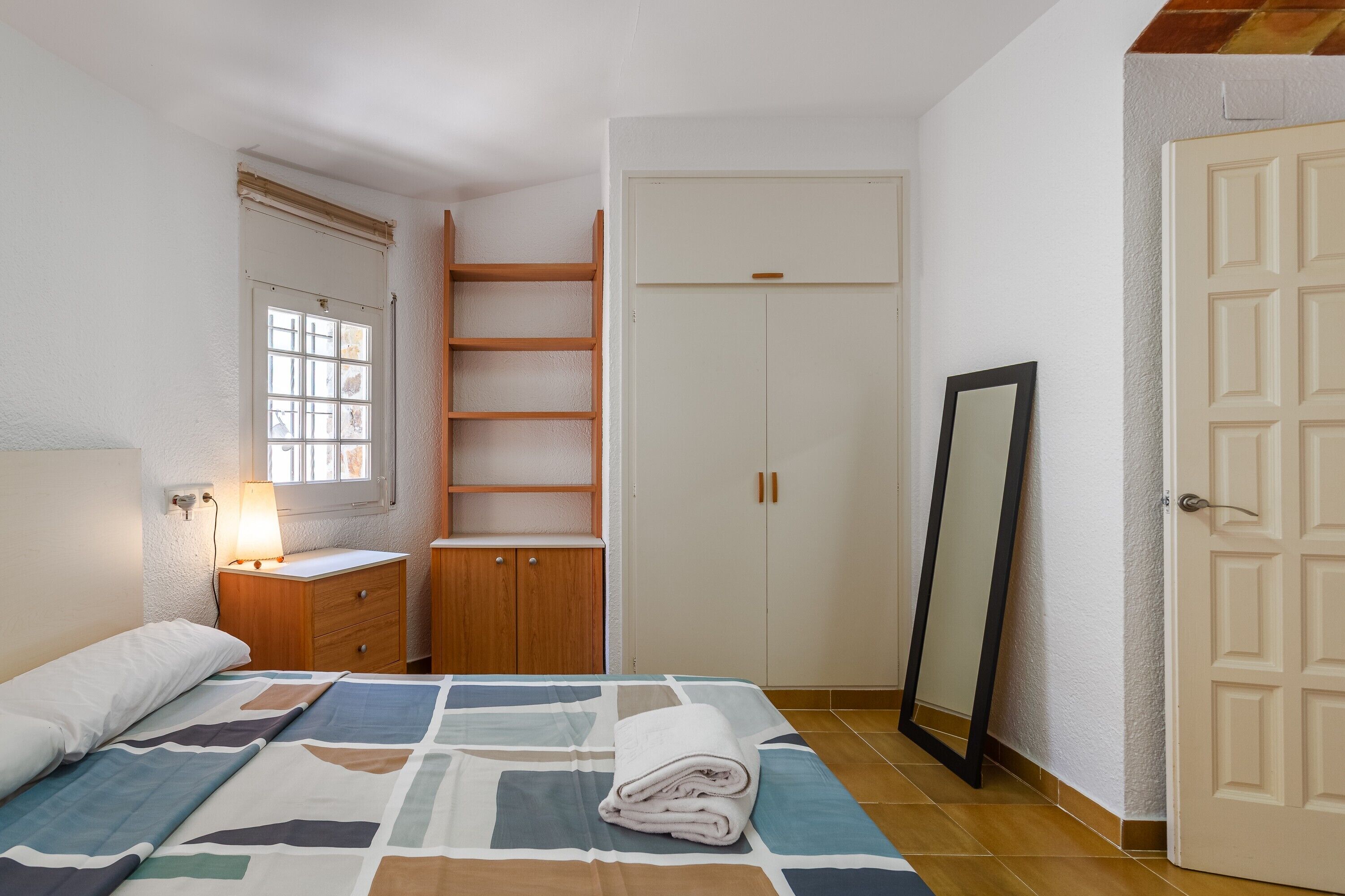 Une chambre à coucher avec un lit, une table de chevet en bois, une armoire, un miroir et une fenêtre.