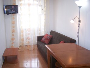 Uma sala de estar com sofá, uma mesinha, televisão fixada na parede e abajur.