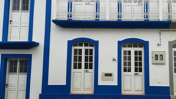 A building with white doors and blue trim, a balcony with white railings, and a small window with a light fixture. - Apartment 'Moradia Machado' with Private Terrace, Balcony and Wi-Fi (Topo Calheta São Jorge Açores)