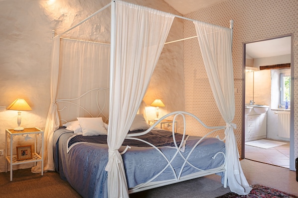 A four-poster canopy bed with a white metal frame, two bedside tables with lamps, a framed picture on one table, and a view into the bathroom through an open door.