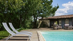 Une terrasse en bois avec des chaises longues, une piscine et un bâtiment rustique doté d’un toit en tuiles.