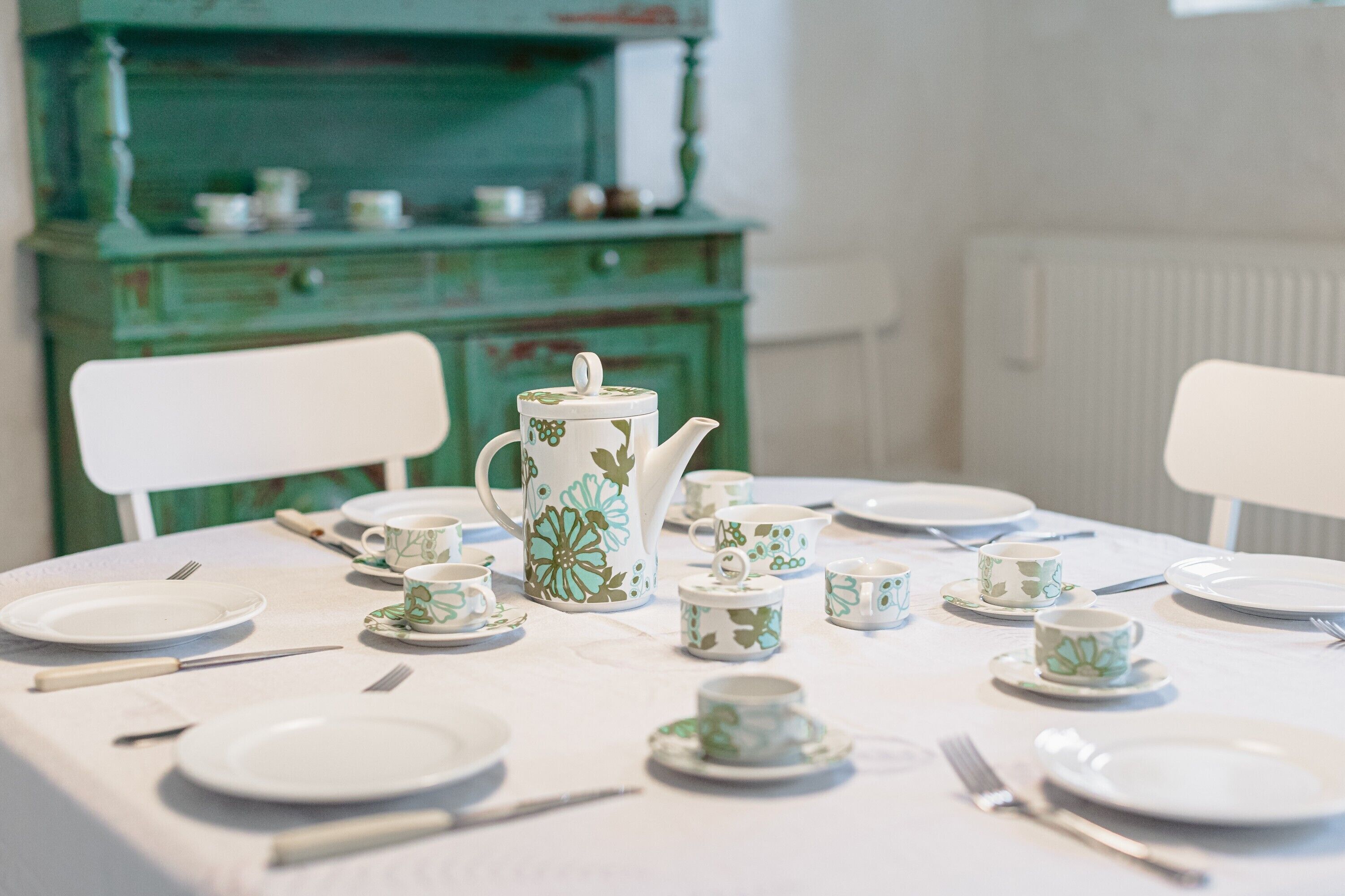 Una mesa dispuesta para una comida, con platos, tazas y una tetera blancos, y un aparador verde al fondo.