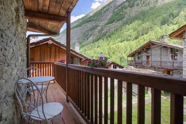 A wooden balcony with a stone wall, a table with chairs, and a view of a mountain.